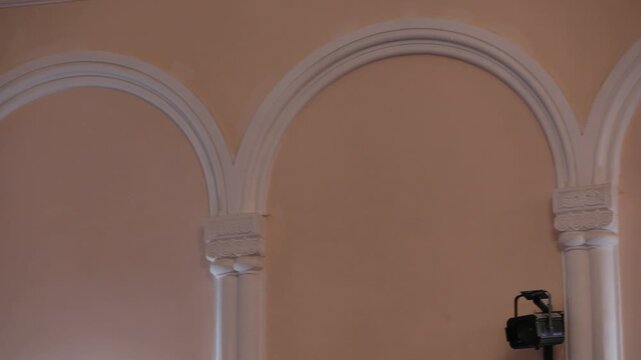 Close-up pan across intricate architectural details inside a classic building. The shot features white plaster moldings, arches, and columns against a warm, elegant peach-colored wall interior.

