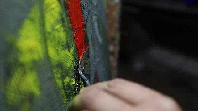 Close-up of artist's hand dipping paintbrush into solvent cup on colorful wooden palette. Detailed shot of creative painting process in art studio environment with oil paints.

