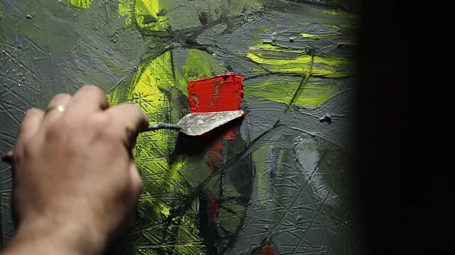 Close-up of artist's hand dipping paintbrush into solvent cup on colorful wooden palette. Detailed shot of creative painting process in art studio environment with oil paints.


