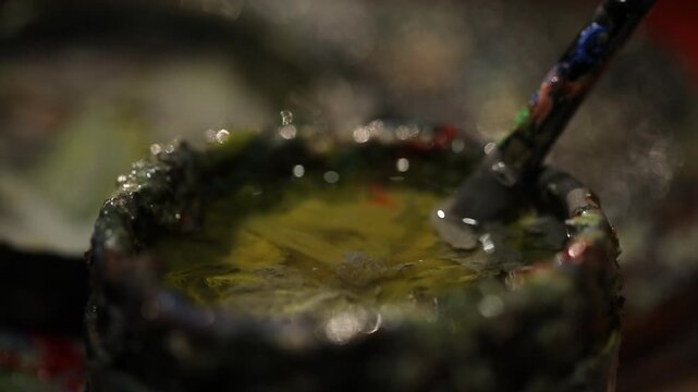 Close-up of artist rinsing paintbrush in small ceramic cup filled with solvent liquid. Bubbles form as brush swirls in the yellow-tinted cleaner on the painter's palette.

