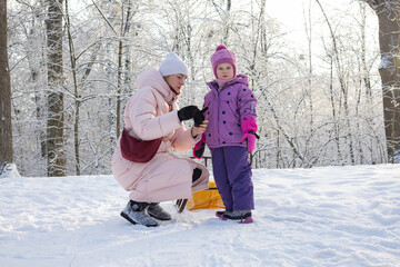 Mother kneeling and adjusting child's gloves before sledding on hill
