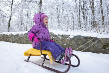 Joyful little girl laughing while mother pulls her on sled through snow covered park in winter