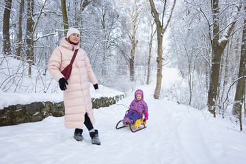 Happy mother pulling toddler daughter on wooden sled through snowy winter forest park during weekend walk