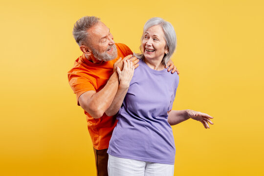 Smiling positive senior couple wearing colorful clothes hugging together on yellow background