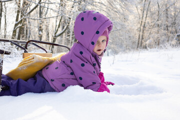Cute little girl in purple winter overalls lying on fresh white snow in sunny forest
