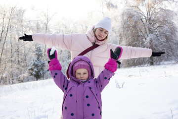Cheerful mother and little daughter posing with open arms in sunny winter forest park