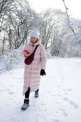 Young woman in long pink coat standing on snowy path among frosted trees in winter park