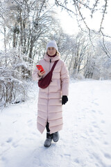 Young woman in pink down jacket checking her smartphone while walking in snowy winter forest
