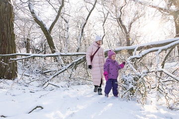 Young mother in pink coat walking with little daughter among snow covered trees in sunny winter park
