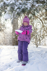 Cute little girl in purple winter overalls playing and crawling in deep white snow in sunny forest