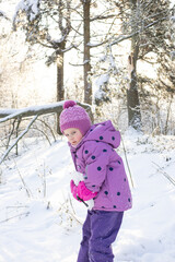 Portrait of a little girl in purple winter suit and pink hat looking at camera in snowy forest