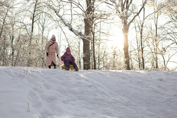 Young mother and little daughter with yellow sled standing on top of snowy hill in winter park