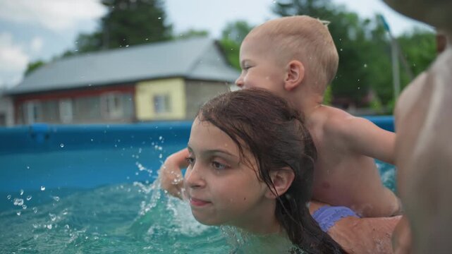 Caucasian Kids Piggybacking In Backyard Pool, Older Sister Carrying Little Brother On Her Back While They Laugh And Splash, Energetic Summer Play With Rustic Homes In Background, Carefree Sibling