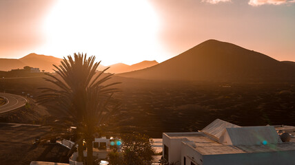 A warm sunset lights La Geria vineyards in Lanzarote, near Timanfaya. A conical mountain rises over lava fields, whitewashed building, palm, and a curving road. © Aerial Film Studio
