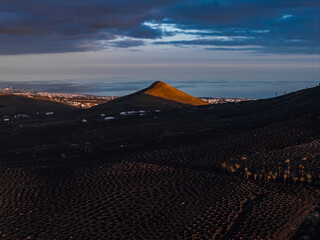 Montana Tinasoria catches sunset light over La Geria vineyards in Lanzarote. Stone zocos, whitewashed houses, and the Atlantic under clouds appear at dusk. © Aerial Film Studio