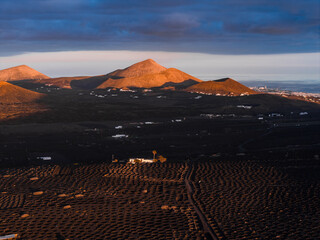 Warm sunset light illuminates La Geria vineyards and volcanic cones in Lanzarote. White farmhouses sit on lava fields, with the Atlantic and Arrecife on the horizon. © Aerial Film Studio