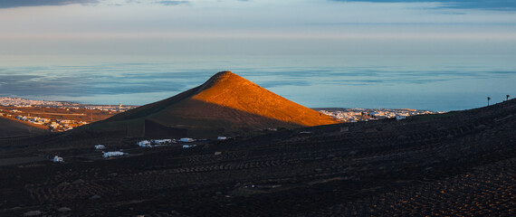 Terraced stone pits and semicircular lava walls frame vines in La Geria on Lanzarote. Whitewashed farmhouses and a coastal town face the Atlantic under soft evening clouds. © Aerial Film Studio