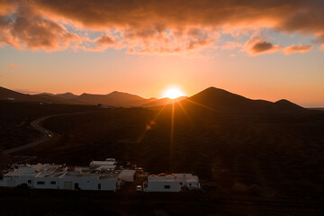 Glowing sunset light bathes Lanzarote as the sun dips behind Timanfaya peaks. Whitewashed bodega sits by a winding road through La Geria vineyards and dark lava fields. © Aerial Film Studio