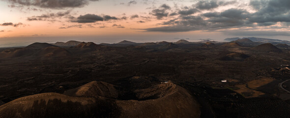 Aerial view at dusk shows Lanzarote badlands with conical volcanoes, ash craters, dark lava fields, and vineyard pits near Timanfaya and La Geria, facing the Atlantic.