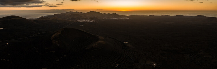 Aerial view at sunset in Lanzarote shows La Geria vineyards, cinder cone, and Timanfaya ridges, with the sun dipping into the Atlantic near Yaiza and Uga. © Aerial Film Studio