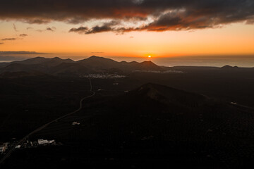 Aerial sunset over Lanzarote shows volcanic cones, La Geria vineyards, Timanfaya lava fields, and winding roads as the sun dips toward the Atlantic under low clouds.