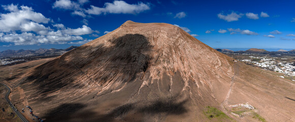 Aerial view shows an ochre volcanic cone near La Geria in Lanzarote, Canary Islands. Midday light, scattered clouds, ridge paths, towns to the right, and a curving road with cars. © Aerial Film Studio