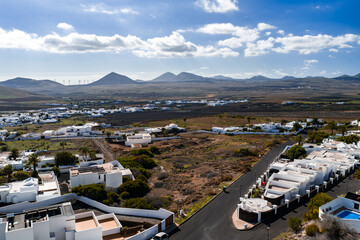Aerial view over Lanzarote shows white houses, dark lava fields, conical volcanoes, La Geria soils, wind turbines, a serpentine road, and terraced villas in late morning light. © Aerial Film Studio
