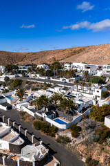 Aerial view shows whitewashed villas, blue pools, palm gardens, and volcanic slopes in Lanzarote, Canary Islands, likely near Teguise or Arrecife, under clear daylight. © Aerial Film Studio