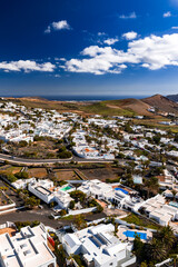 Aerial view shows whitewashed village in Lanzarote, Canary Islands, with cubic houses, pools, palm trees, terraced volcanic fields, and Atlantic Ocean under clouded sky. © Aerial Film Studio