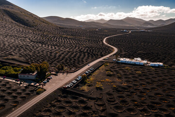 Aerial view shows La Geria vineyards with crescent zocos, a winding road, whitewashed buildings, and parked cars. Volcanic cones rise on the horizon in Lanzarote. © Aerial Film Studio