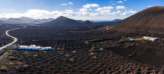 Aerial view shows La Geria volcanic vineyards on Lanzarote, Canary Islands, with circular hollows, stone windbreaks, whitewashed farms, road, and Timanfaya cones at midday. © Aerial Film Studio