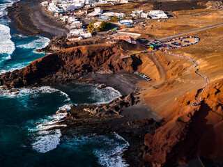 Aerial scene shows El Golfo cliffs, black sand cove, green lagoon, and whitewashed village on Lanzarote. Waves strike lava outcrops near a boardwalk, road, and car park at late afternoon. © Aerial Film Studio