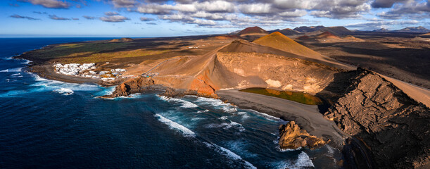Aerial view shows Charco Verde and El Golfo on Lanzarote. A crater wall meets a black sand beach and emerald lake. Timanfaya cones line the horizon under clouds. © Aerial Film Studio