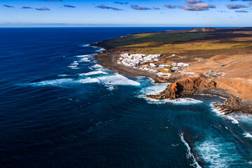 Aerial view shows El Golfo beside the Atlantic, black lava fields, ochre cliffs, and the green Charco de los Clicos near Timanfaya under midday light. © Aerial Film Studio
