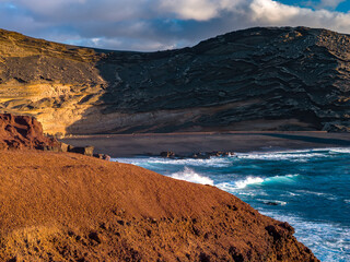 Turquoise surf hits a black sand beach at El Golfo near Charco de los Clicos, Lanzarote. Rust red lava rock, layered tuff and basalt, and a slim boardwalk appear in late light.