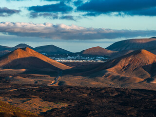 Rust cinder cones and lava fields frame a winding road to whitewashed buildings in Lanzarote near La Geria and Timanfaya, with long shadows and layered clouds. © Aerial Film Studio