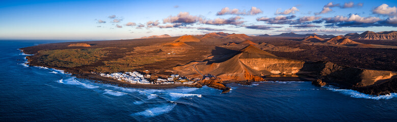 Aerial view of Lanzarote southwest coast at golden hour, Atlantic surf meets dark volcanic shore, Timanfaya cones and ash fields rise, El Golfo road runs by a whitewashed village © Aerial Film Studio