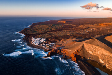 Aerial scene shows El Golfo coast in Lanzarote, Timanfaya cliffs, lava fields, white houses, surf coves, and a road on cinder slopes in warm late afternoon light. © Aerial Film Studio