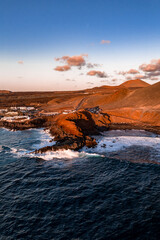 Aerial scene of Lanzarote west coast shows El Golfo and Timanfaya, Atlantic waves on black cliffs, rust fields, coastal road, white buildings, and a conical volcanic hill. © Aerial Film Studio