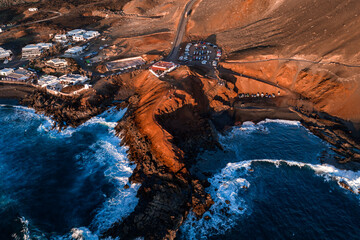 Aerial view shows Lanzarote volcanic coast near El Golfo and Charco de los Clicos. Lava cliffs meet turquoise surf, roads and paths trace slopes in late afternoon light. © Aerial Film Studio
