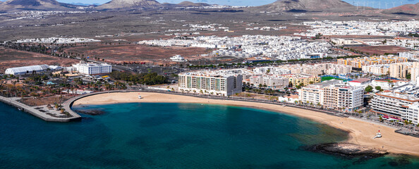 Fototapeta premium Aerial view of Arrecife, Lanzarote, shows Playa del Reducto, turquoise water, promenade, mid rise hotels, white buildings, volcanic hills, wind turbines, bright daylight.