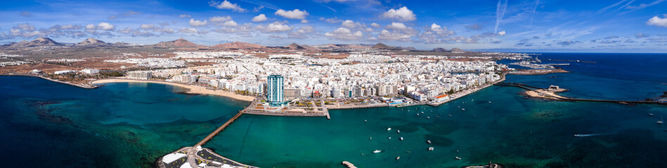 Aerial view of Arrecife, Lanzarote shows whitewashed city, marina, breakwaters, small boats, a modern high rise, and Castillo de San Gabriel causeway under daylight skies. © Aerial Film Studio