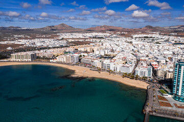 Aerial view of Arrecife, Lanzarote shows Playa del Reducto, promenade, mid rise hotels, volcanic cones, wind turbines, and teal bay under bright midday light and clouds.