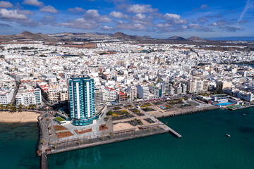Aerial view of Arrecife, Lanzarote shows Gran Hotel and Spa by a white grid, volcanic hills, turquoise Atlantic, promenade, small beach, piers, plazas, and palm walkways at midday. © Aerial Film Studio