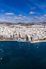 Aerial view of Arrecife, Lanzarote, shows whitewashed buildings by a calm bay with small boats, volcanic hills beyond, and a waterfront promenade under bright midday light. © Aerial Film Studio