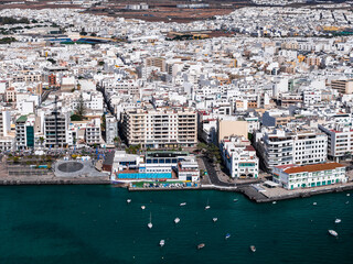 Aerial view of Arrecife, Lanzarote, Spain, shows whitewashed buildings, turquoise waterfront, seafront promenade, marinas, anchored boats, and a sunlit mid rise skyline. © Aerial Film Studio