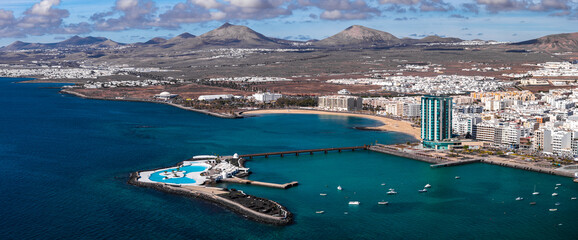 Aerial view of Arrecife, Lanzarote shows Playa del Reducto, Islote de Fermina pools, Arrecife Gran Hotel, fishing boats, a pedestrian pier, and volcanic cones under clouds.