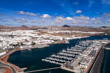 Aerial view of Puerto Deportivo Marina Lanzarote and commercial port in Arrecife, with sailboats, white buildings, breakwaters, volcanic cones, clear skies, and turquoise water.