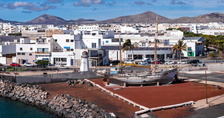 A small lighthouse stands by a preserved wooden sailing ship on land near rocky shore. Arrecife, Lanzarote, Canary Islands, shows wind turbines and volcanic mountains. © Aerial Film Studio