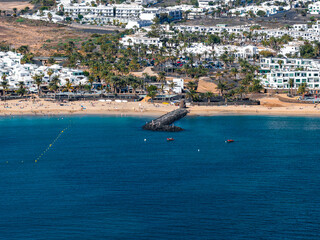 Sandy beach in Lanzarote shows palm trees, white resorts, and a stone jetty. Swimmers and boats are present, with a buoy line marking safe bathing in bright daylight. © Aerial Film Studio